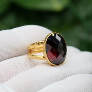 May include: A gold ring featuring a large, faceted, oval-shaped, dark red gemstone. The ring has a double-band design and is set against a white background with a blurred green backdrop. The gemstone is approximately 1 inch in length.
