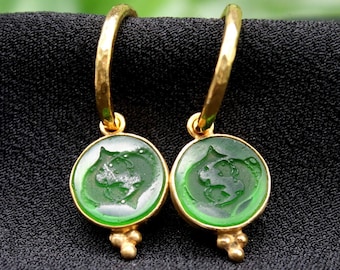 May include: A pair of gold-colored hoop earrings with green, circular pendants. Each pendant features a detailed, embossed design. The earrings are displayed on a black surface, highlighting their intricate details and color.