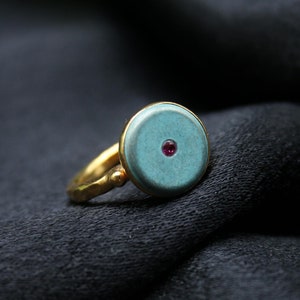 May include: A gold ring with a turquoise blue circular stone set in the center. The stone has a small red gemstone in the center.