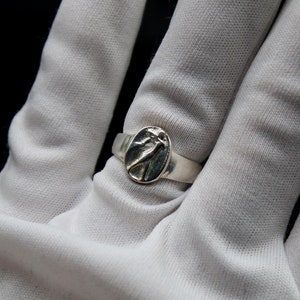 Pu&ograve; includere: Un anello d'argento con un design ovale che presenta un uccello stilizzato. L'anello &egrave; indossato su una mano guantata di bianco.