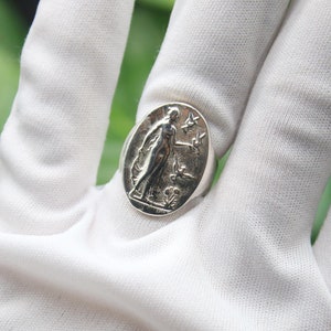 Può includere: Un anello d'argento con un design ovale che presenta una donna in piedi con un uccello in mano. L'anello è su una superficie bianca.