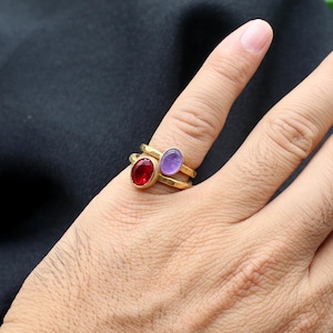 May include: Two gold rings with oval-shaped gemstones. The top ring has a purple gemstone and the bottom ring has a red gemstone.