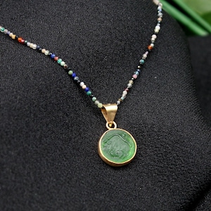 May include: A green gemstone pendant with a gold setting hangs from a multi-colored beaded necklace. The necklace is on a black fabric background.