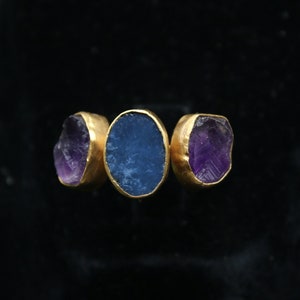 May include: A gold ring with three stones. The center stone is a blue oval, and the two stones on either side are purple.