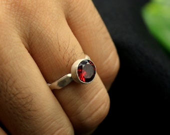May include: A silver ring with a red gemstone. The ring is on a person's finger.