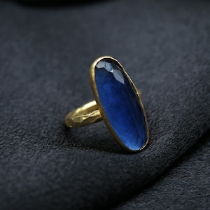 May include: A gold-toned ring with a long, oval-shaped, deep blue gemstone. The gemstone is set in a gold bezel and has a faceted surface. The ring's band has a textured, hammered appearance, adding a rustic touch to the jewelry.