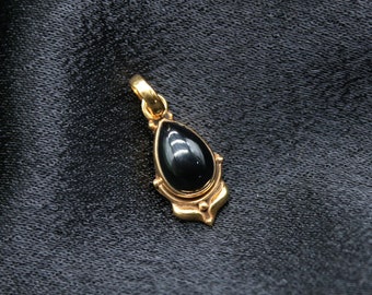 May include: A gold-colored pendant with a teardrop-shaped black stone. The pendant has a decorative gold frame and a loop for a chain. The jewelry is displayed on a dark, textured fabric.