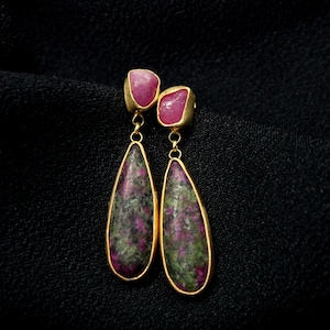 May include: A pair of gold-coloured earrings with a teardrop design. The earrings feature a combination of pink and green stones, with a smaller pink stone at the top. The earrings are set against a black background.