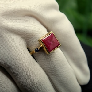 May include: A gold ring with a large, square, red gemstone and a smaller, round, blue gemstone.