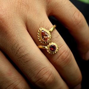 May include: A gold ring with two teardrop-shaped stones. The larger stone is a light orange colour, and the smaller stone is a deep red.