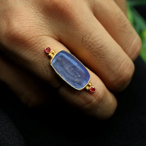 May include: A gold ring with a blue stone set in a rectangular frame. The stone has a carved design and is surrounded by two small red stones.