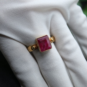 May include: A gold ring with a large, square, red ruby gemstone set in the center. The ring has small gold accents on the band.