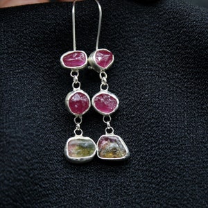 May include: Silver dangle earrings with three pink and green gemstones. The top two gemstones are pink and the bottom gemstone is green, pink, and yellow.