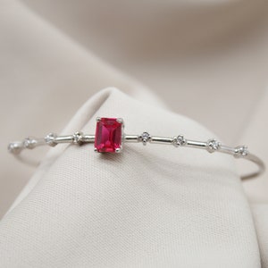 Könnte beinhalten: Ein silbernes Armband mit einem roten Edelstein und kleinen klaren Edelsteinen.