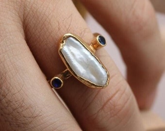May include: A gold ring featuring a large white baroque pearl and two small blue gemstones. The ring is worn on a finger.