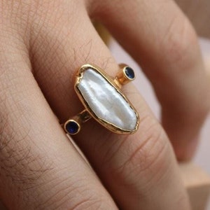May include: A gold ring with a large white baroque pearl and two small blue gemstones. The ring is on a finger.