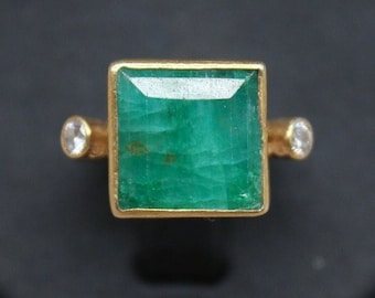 Op de afbeelding: Een gouden ring met een grote, vierkante smaragd edelsteen en twee kleinere diamanten aan de zijkanten.