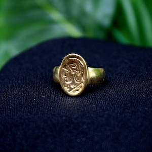 Pode incluir: Um anel de ouro com um camafeu de uma pessoa com um capacete. O anel est&aacute; sobre um fundo de tecido preto.
