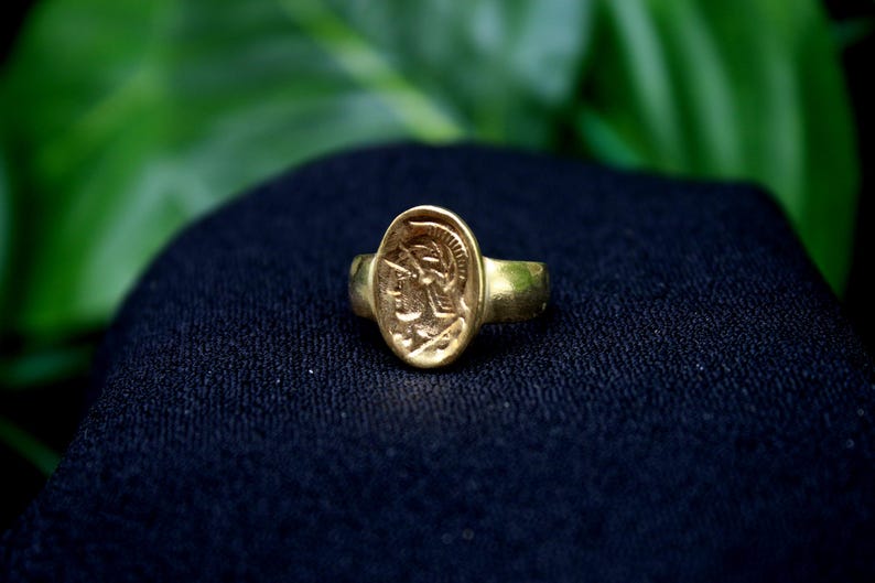 Pode incluir: Um anel de ouro com um camafeu de uma pessoa com um capacete. O anel est&aacute; sobre um fundo de tecido preto.