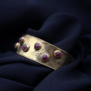 May include: A gold cuff bracelet with a hammered texture and red gemstones set into the metal.