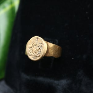 May include: A gold signet ring with a detailed engraved design of a mythical creature. The ring is set on a black velvet background.