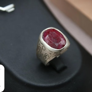 May include: A silver ring with an oval-shaped red gemstone set in the center. The ring has an intricate design around the gemstone.