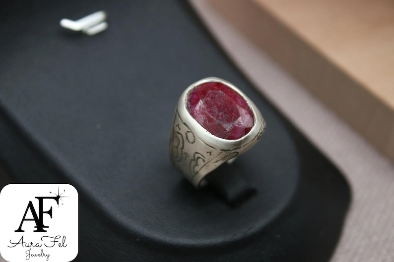 May include: A silver ring with an oval-shaped red gemstone set in the center. The ring has an intricate design around the gemstone.