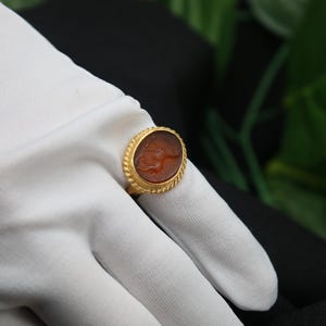 May include: A gold ring with an oval-shaped carnelian stone featuring a carved design. The ring is set on a white surface.