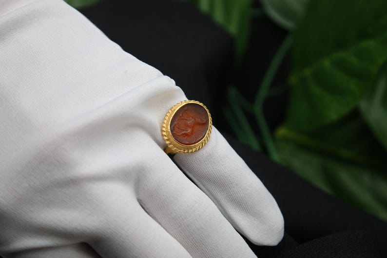 May include: A gold ring with an oval-shaped carnelian stone featuring a carved design. The ring is set on a white surface.