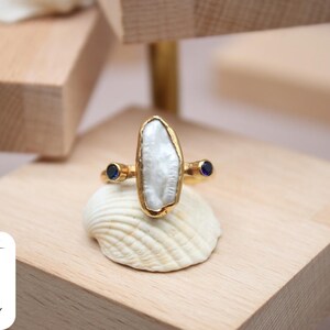 May include: A gold ring with a large, white, irregular-shaped pearl and two small, blue sapphire accents.