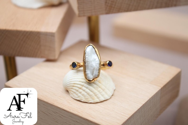 May include: A gold ring with a large, white, irregular-shaped pearl and two small, blue sapphire accents.
