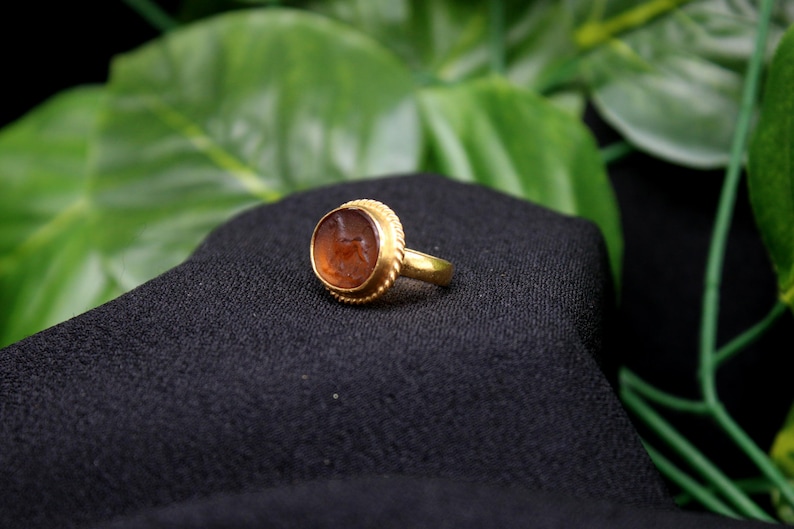 May include: A gold ring with a brown oval-shaped stone set in a gold bezel. The stone has a detailed carving on its surface.