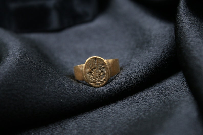 May include: A gold ring with an oval-shaped design featuring a detailed image of a person holding a sword. The ring is on a dark gray fabric background.