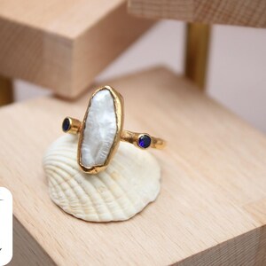 May include: A gold ring with a large white pearl-like stone and two small blue stones. The ring is sitting on a white seashell.