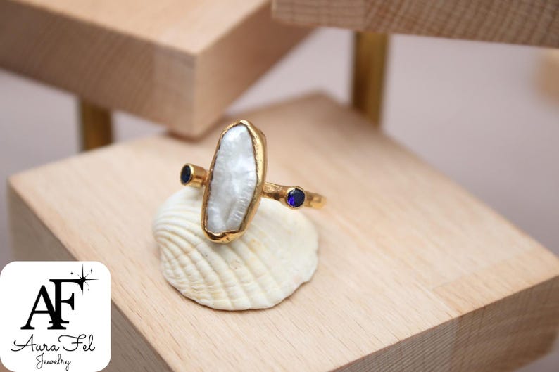 May include: A gold ring with a large white pearl-like stone and two small blue stones. The ring is sitting on a white seashell.