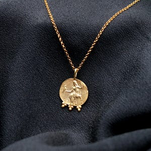 May include: A gold chain necklace with a round pendant featuring a detailed image of a woman riding a horse. The pendant has small gold beads hanging from the bottom.