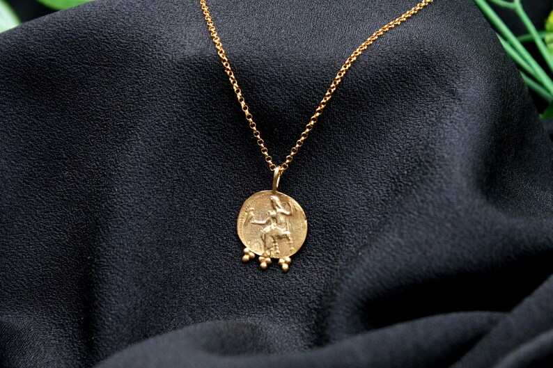 May include: A gold chain necklace with a round pendant featuring a detailed image of a woman riding a horse. The pendant has small gold beads hanging from the bottom.