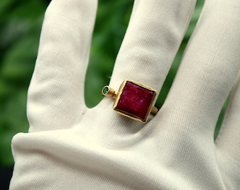 Könnte beinhalten: Ein goldener Ring mit einem quadratischen roten Edelstein und einem kleinen grünen Edelstein an der Seite.