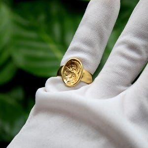 Pode incluir: Um anel de ouro com um design oval que apresenta uma imagem detalhada da cabe&ccedil;a de uma pessoa de perfil. O anel &eacute; usado em um dedo enluvado de branco.
