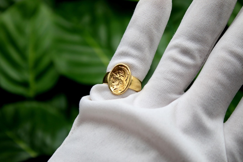 Pode incluir: Um anel de ouro com um design oval que apresenta uma imagem detalhada da cabe&ccedil;a de uma pessoa de perfil. O anel &eacute; usado em um dedo enluvado de branco.