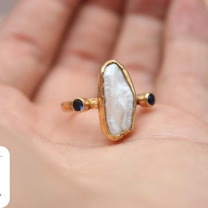 May include: Gold ring with a large white pearl-like stone and two small blue gemstones.