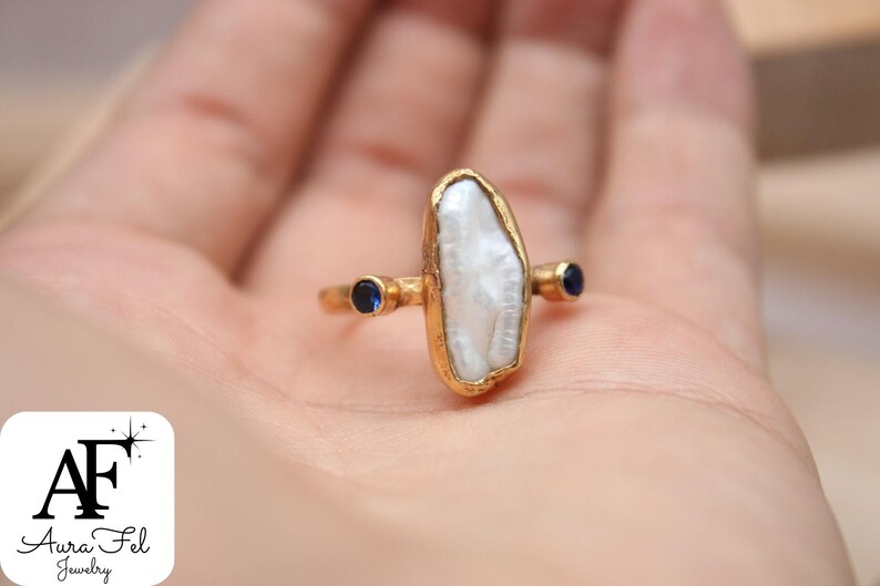 May include: Gold ring with a large white pearl-like stone and two small blue gemstones.