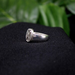 Pu&ograve; includere: Un anello d'argento con un design ovale testurizzato. L'anello &egrave; su uno sfondo di tessuto nero.