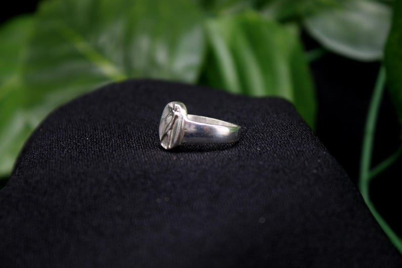 Pu&ograve; includere: Un anello d'argento con un design ovale testurizzato. L'anello &egrave; su uno sfondo di tessuto nero.