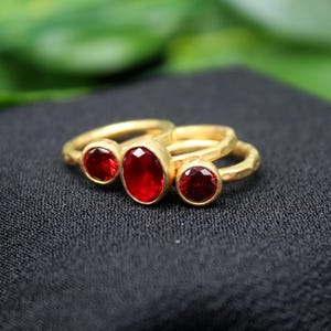 May include: Three gold rings with red oval-shaped gemstones. The rings are stacked on top of each other.