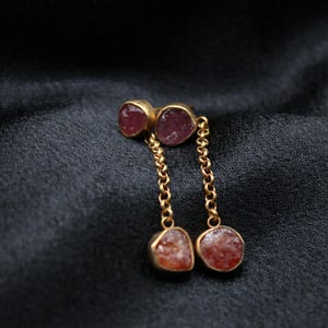 May include: A pair of gold earrings with pink and orange gemstone drops. The earrings feature a delicate chain connecting the top and bottom stones.