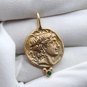 May include: A gold-colored pendant featuring a profile of a person with laurel leaves, reminiscent of ancient Greek or Roman art. The pendant has a small green gemstone at the bottom and a loop at the top for a chain.