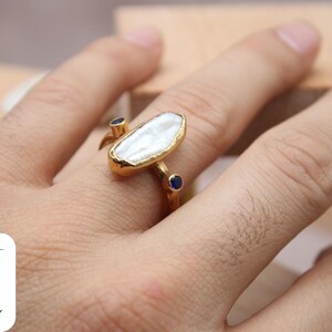May include: Gold ring with a large white baroque pearl and two small blue sapphires. The ring is on a person's finger.