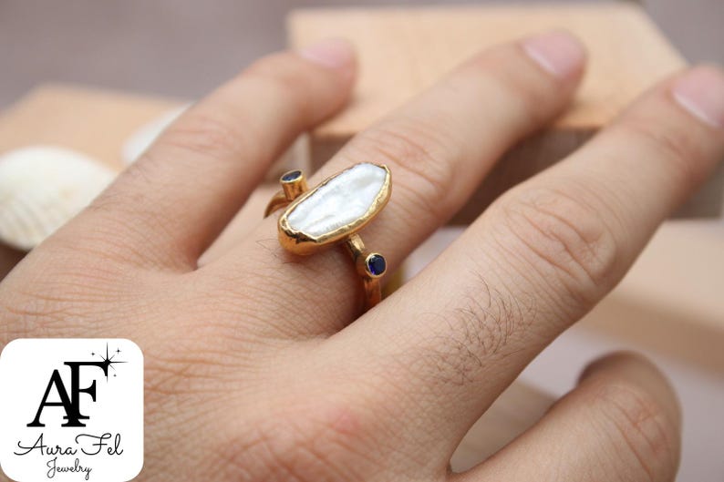 May include: Gold ring with a large white baroque pearl and two small blue sapphires. The ring is on a person's finger.