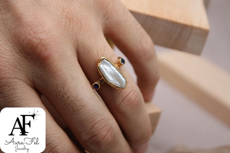 May include: A gold ring with a large white baroque pearl and two small blue gemstones. The ring is on a finger.